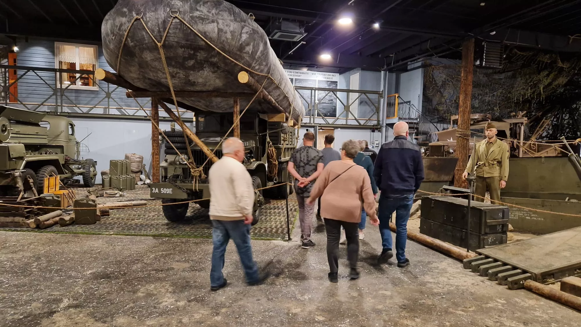 Mensen bezoeken een museum over de Tweede Wereldoorlog en bekijken spullen zoals een legerauto en een legerboot